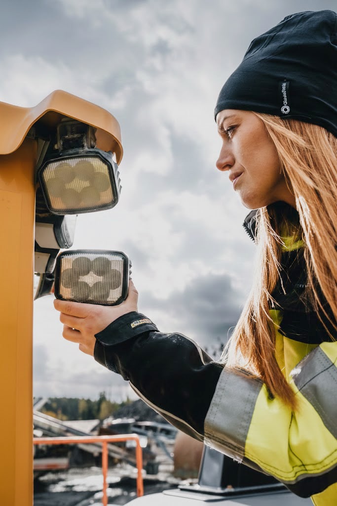 Kvinna i varselväst och mössa vid strålkastare på entreprenadmaskin — employer branding-foto av Grafikbyrån