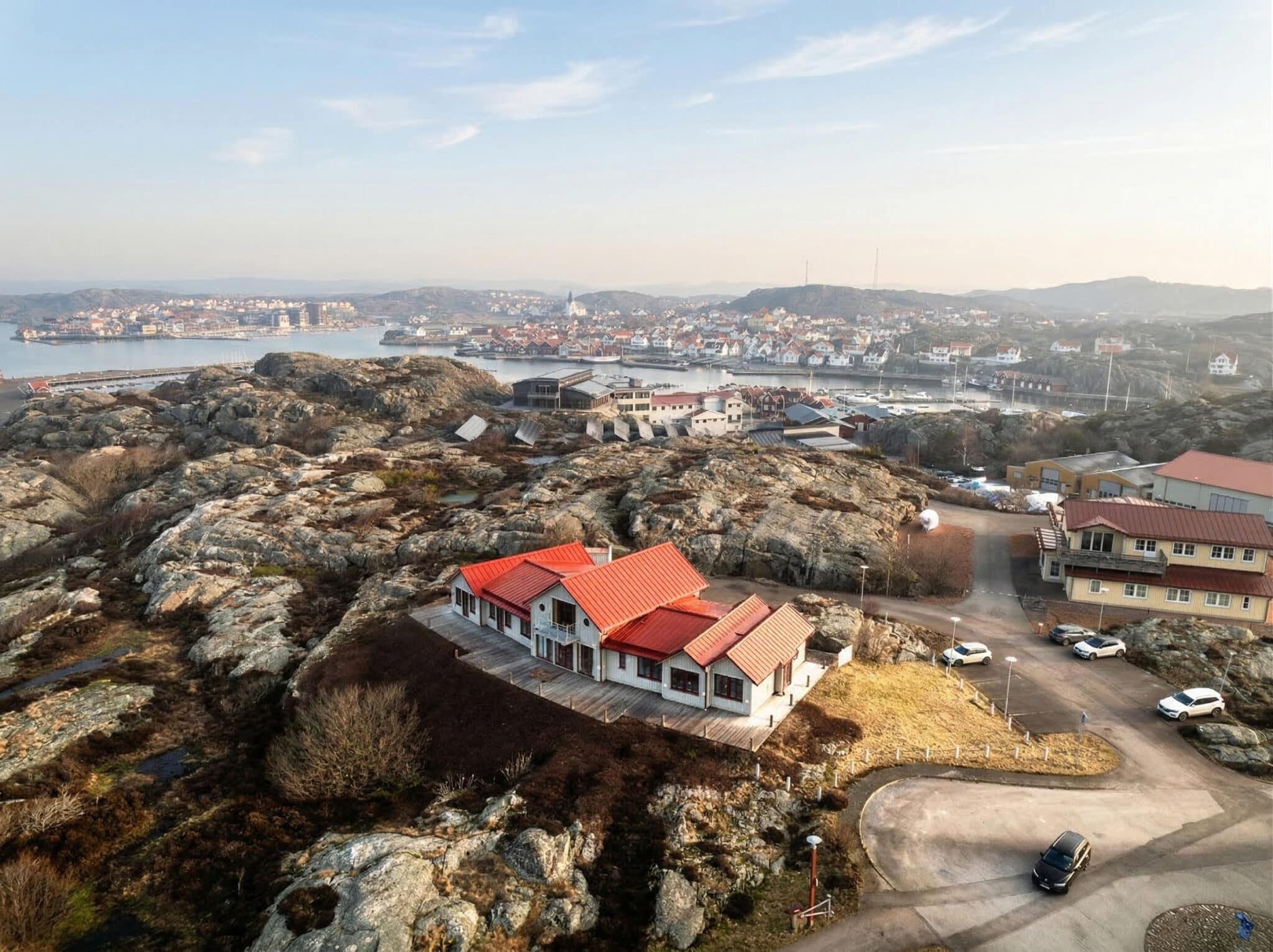 Drönarbild över hus med rött tak i bohuslänsk skärgård med hamn och samhälle i bakgrunden